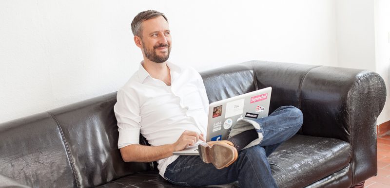Ein Mann sitzt mit einem Laptop auf dem Sofa und lacht zur Seite