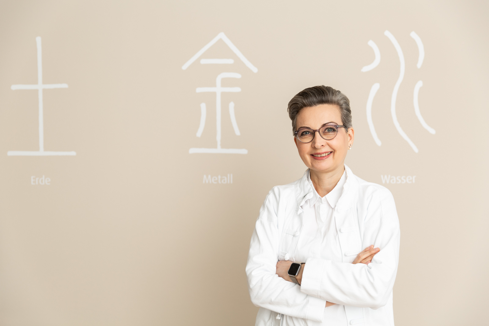 Portrait von Dr. med. Nicola Winckler-Schumann. Sie steht vor einer Wand mit chinesischen Zeichen für die Elemente.